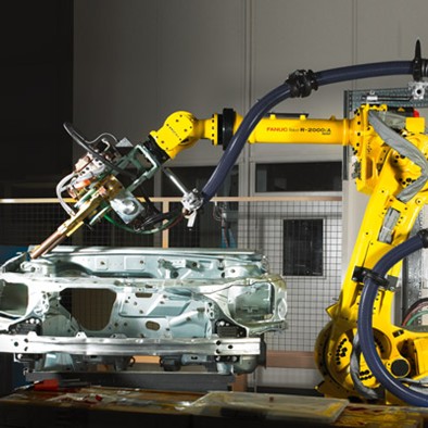 Brazo robótico industrial FANUC equipado con herramientas de ensamblaje trabaja sobre la carrocería de un vehículo en un entorno de fábrica automatizada. El robot está conectado a sistemas de control que optimizan el movimiento mediante tecnología de aprendizaje automático para reducir vibraciones y mejorar la precisión.