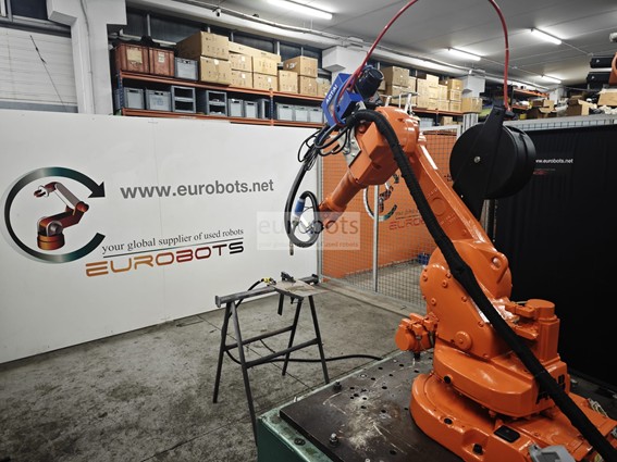 Sistema de soldadura robótica MIG/MAG con brazo robótico industrial naranja trabajando sobre una mesa metálica en un taller, conectado a equipo de soldadura automatizado; al fondo se observan estanterías con cajas y un panel con el logo de Eurobots, representando la automatización industrial para mejorar la productividad y la calidad.