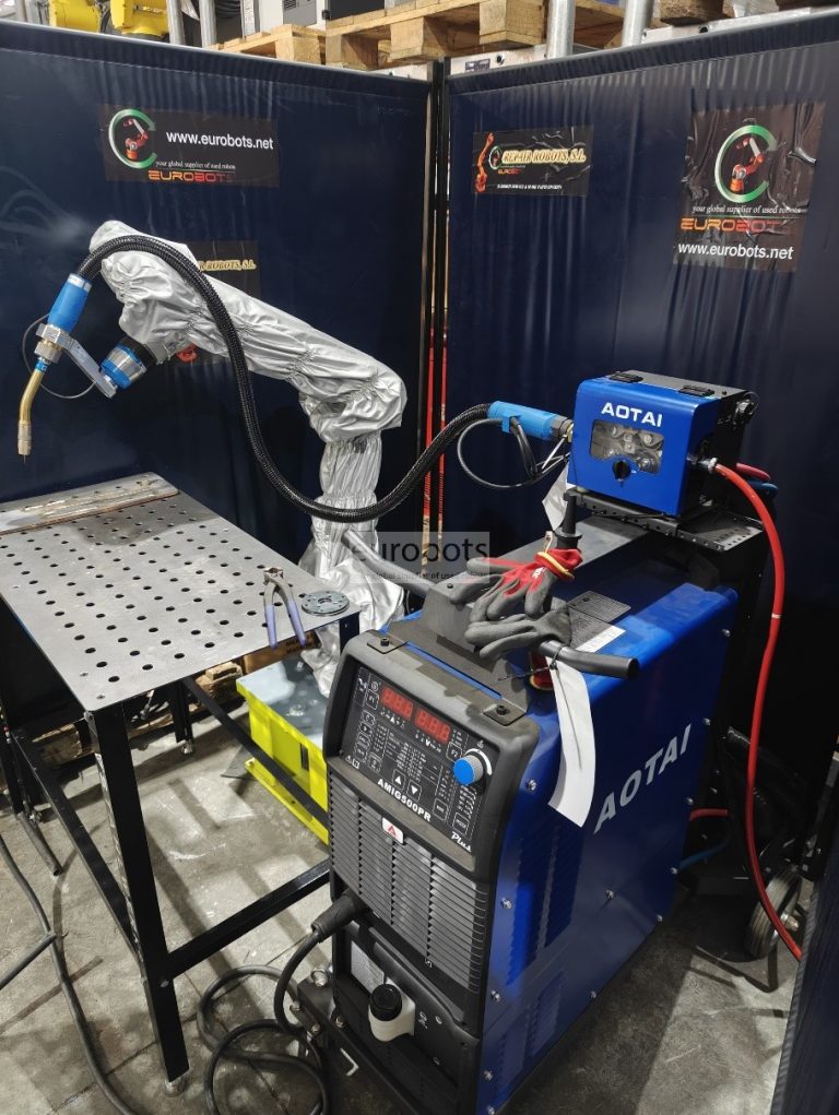 Celda de soldadura robótica en entorno industrial con equipo de soldadura AOTAI conectado a un brazo robótico cubierto con funda protectora. El sistema incluye mesa metálica de trabajo perforada y cables de conexión, diseñado para procesos de soldadura automatizada que detectan variaciones y ajustan parámetros en tiempo real para mejorar la precisión.